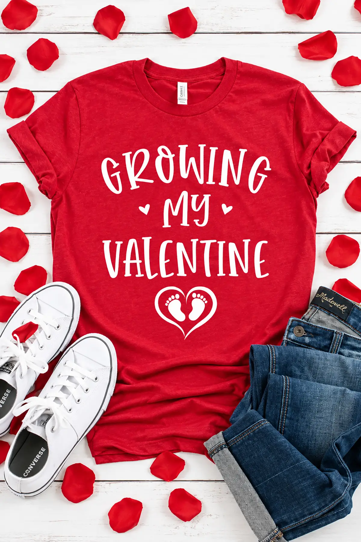 Red "Growing My Valentine" pregnancy announcement T-shirt with baby footprints, styled with jeans, sneakers, and rose petals.