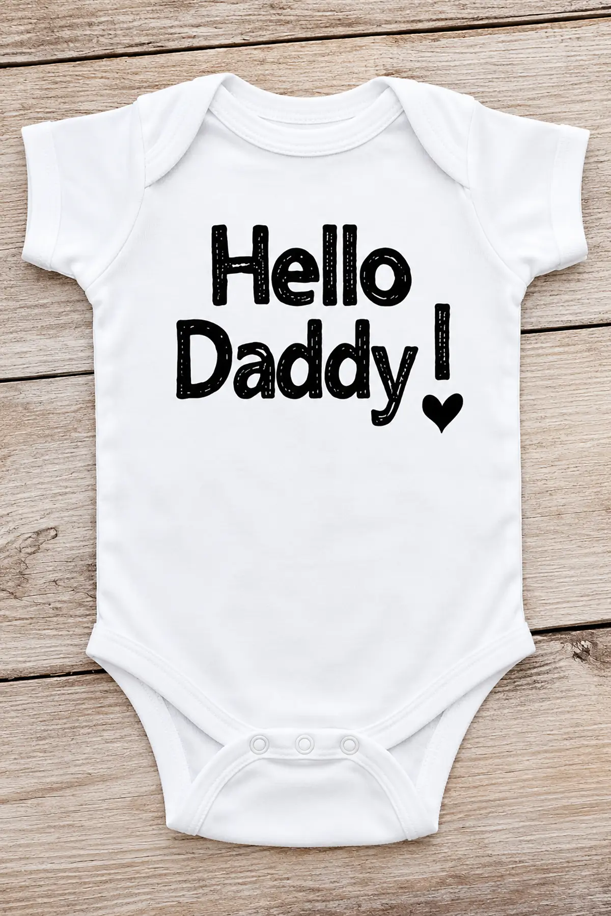 White baby onesie featuring "Hello Daddy!" in black lettering and a heart, displayed on rustic wooden planks.