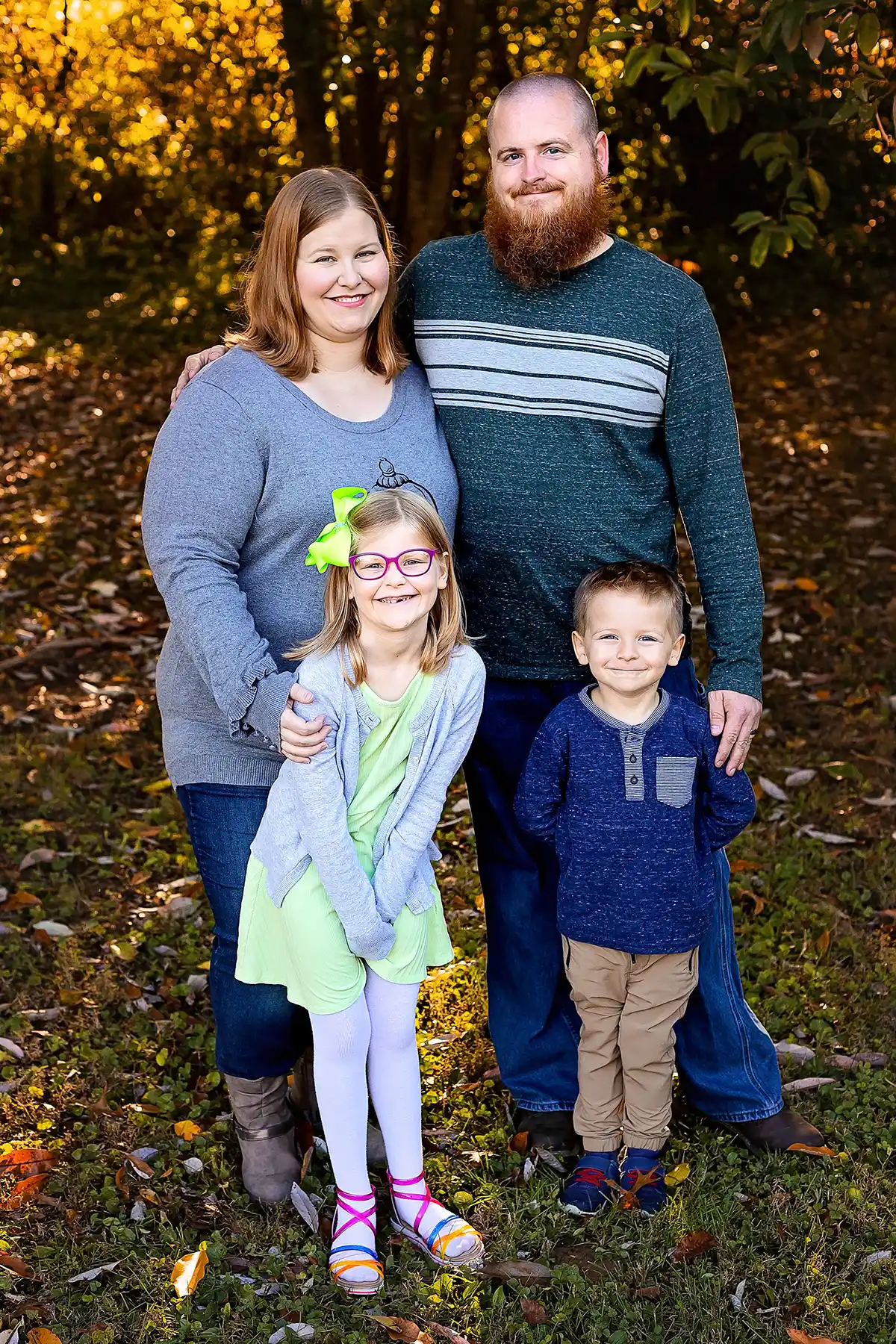 Family of four posing for an outdoor portrait in autumn, with parents standing behind their smiling daughter and son.