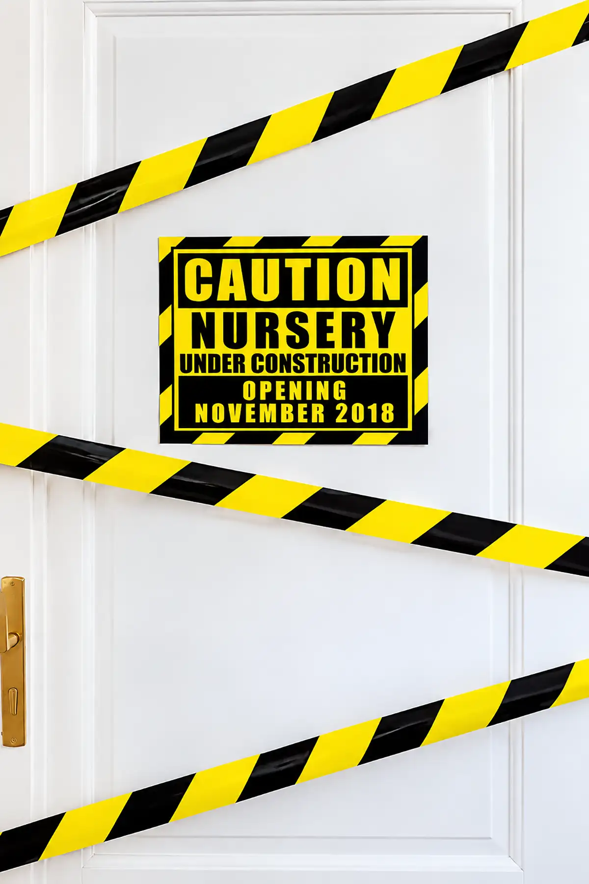 White door decorated with yellow and black caution tape and a "Nursery Under Construction" pregnancy announcement sign.