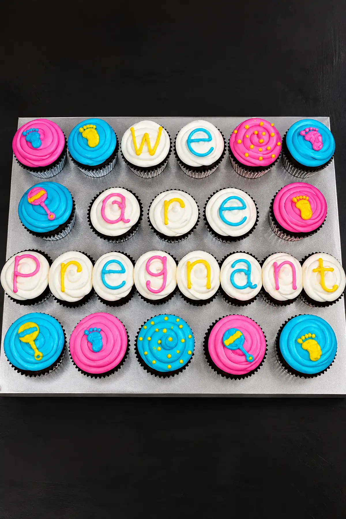 Pink, blue, and white cupcakes decorated with letters spelling "We Are Pregnant" and colorful baby-themed designs.