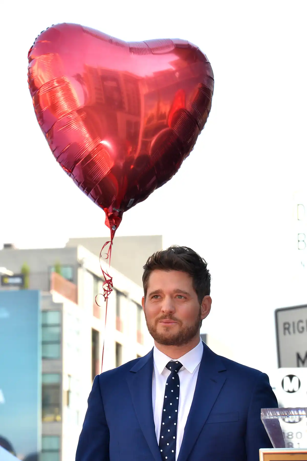 Michael Buble in a navy suit standing beneath a large red heart-shaped balloon, illustrating a romantic singing telegram pregnancy reveal.