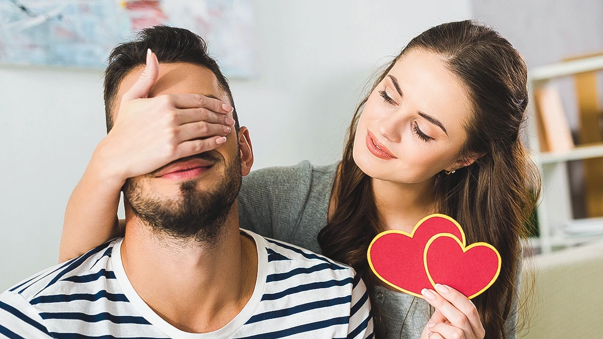 Woman covering her husband's eyes while holding two red hearts, preparing a romantic Valentine's Day pregnancy announcement surprise.