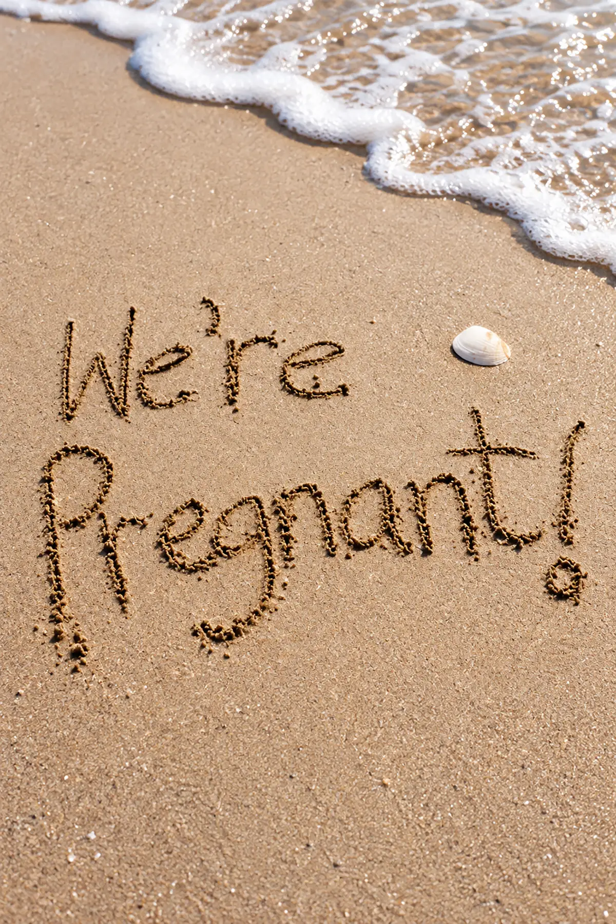 "We're Pregnant!" handwritten in golden beach sand beside a seashell, with ocean waves washing onto the shore.